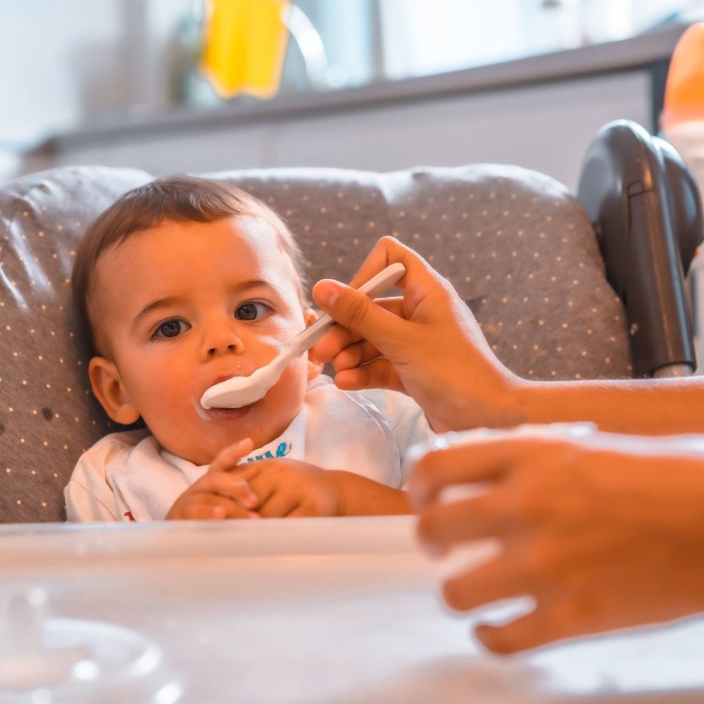 Young Caucasian mother feeding her son a yogurt while sitting in the highchair. babysitting course